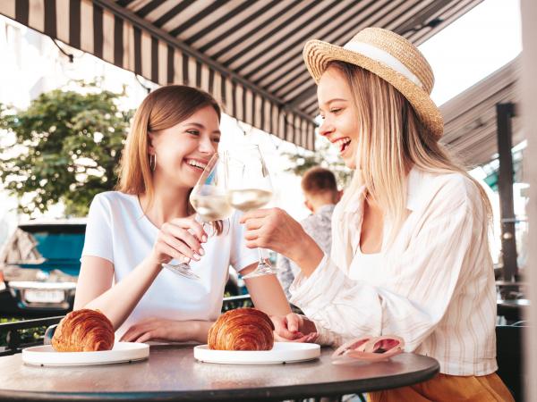dwie mlode piekne usmiechniete kobiety hipster w modnych letnich ubraniachbeztroskie kobiety pozuja na werandzie kawiarni na ulicypozytywne modele pijace biale winocieszacy sie na wakacjachjedzenie rogalika
